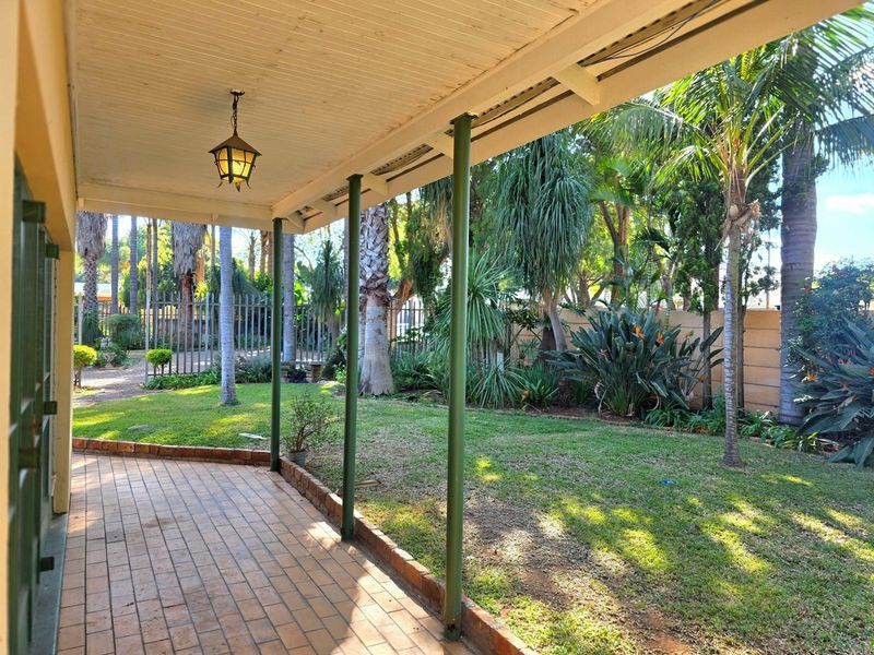 A covered patio area featuring brick paving and metal support columns, overlooking a lush, established garden with mature trees and perimeter fencing.