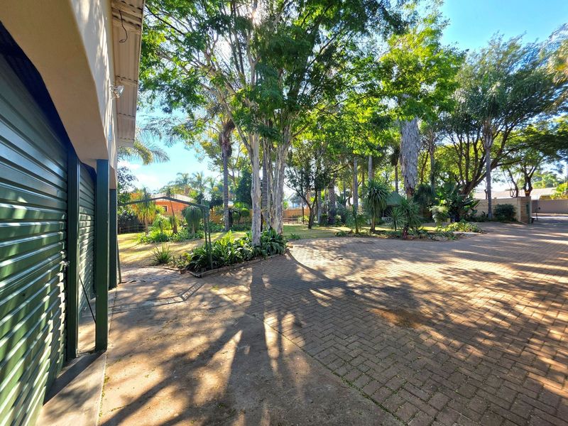Spacious rear exterior area featuring a large paved driveway, mature trees, and established garden landscaping adjacent to the garage.