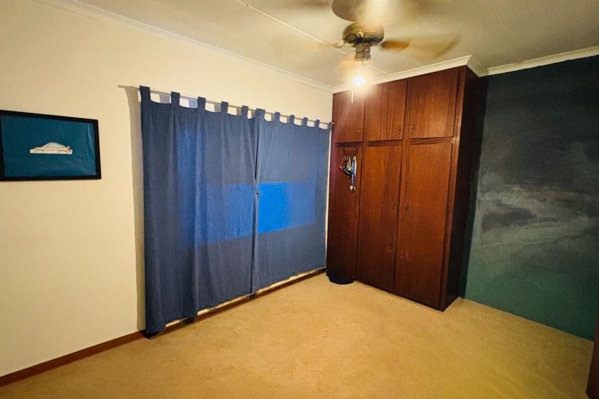 A bedroom featuring built-in wooden cupboards, a ceiling fan, and carpeted flooring, currently presenting with visible wall staining and dated finishes.