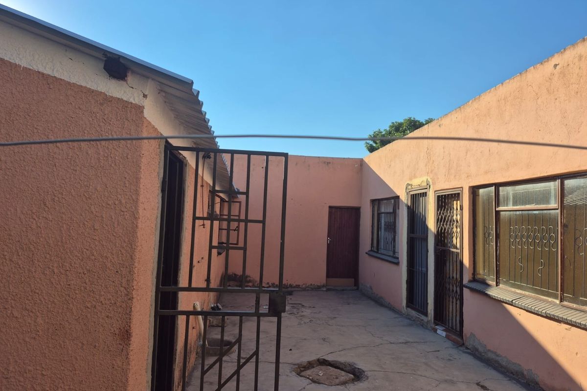 A rear courtyard area featuring concrete paving, security gates, and metal window frames, showing signs of wear and requiring maintenance.
