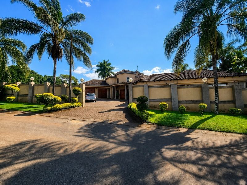 A well-maintained front elevation featuring a paved driveway, secure perimeter walling, double garage, and established landscaped gardens under a clear blue sky.