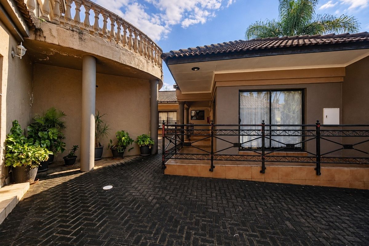 A paved rear courtyard area featuring a raised patio with wrought iron railings, concrete columns, and a balcony with a stone balustrade.