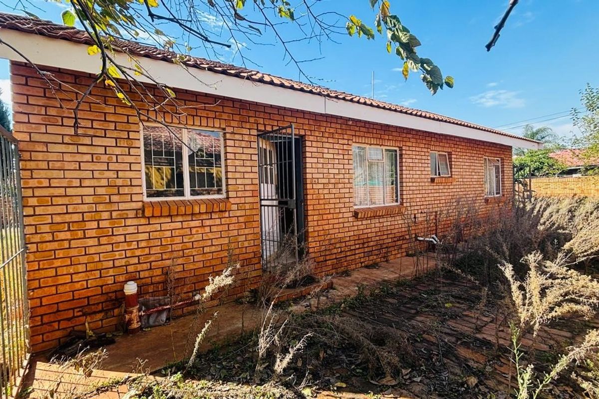 Rear view of a face brick house featuring a tiled roof, metal window frames, and a paved walkway surrounded by an overgrown garden.