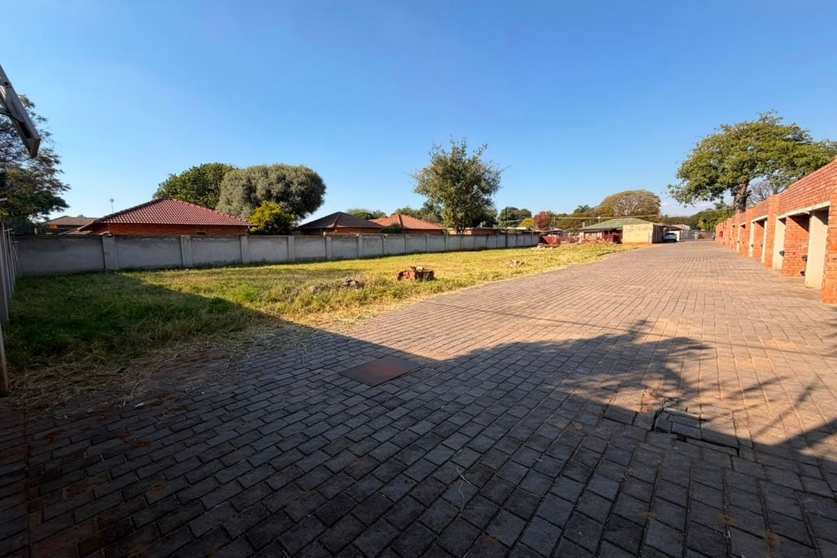 A spacious rear exterior area featuring extensive paved driveway space, a perimeter boundary wall, and an adjacent brick building structure.