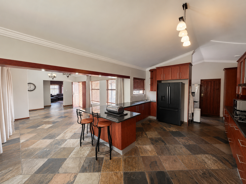 Spacious open-plan kitchen featuring wooden cabinetry, granite countertops, and a central island, seamlessly flowing into the adjacent living area with tiled flooring throughout.