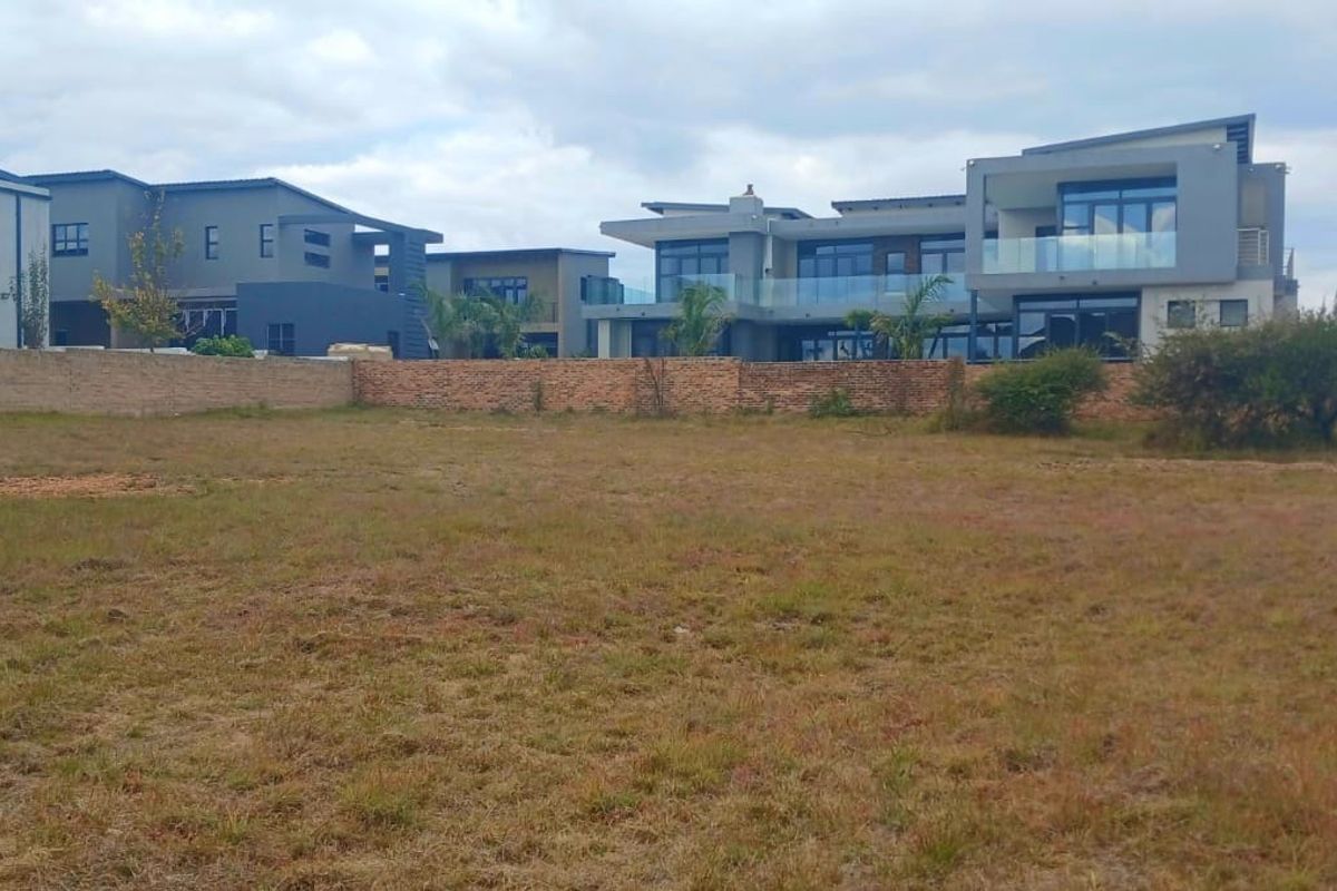 A view of a vacant residential plot featuring a brick boundary wall with established modern homes visible in the background.