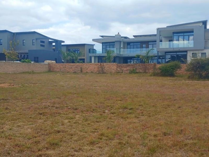 A view of a vacant residential plot featuring a brick boundary wall with established modern homes visible in the background.
