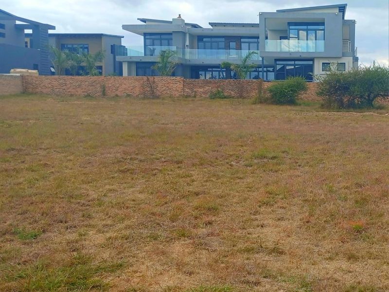 A view of a vacant residential plot with a brick boundary wall, overlooking modern multi-story homes in the background.