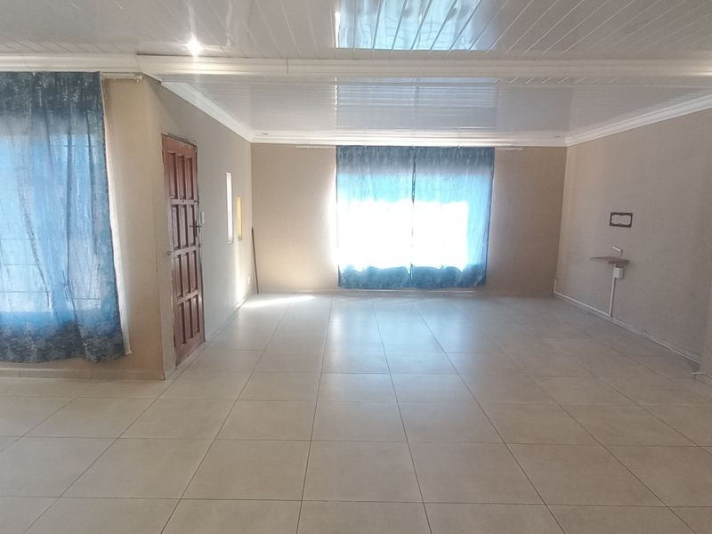 A spacious, empty living area featuring neutral tiled flooring, glossy ceiling panels with recessed lighting, and large windows allowing for ample natural light.