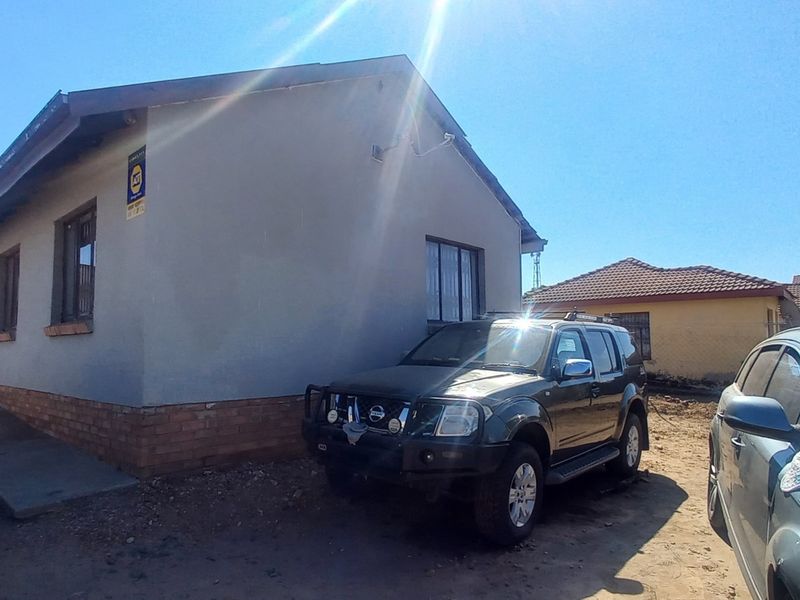 Front elevation of a single-story residential house featuring a brick plinth and basic exterior finishes, situated on an unpaved lot.