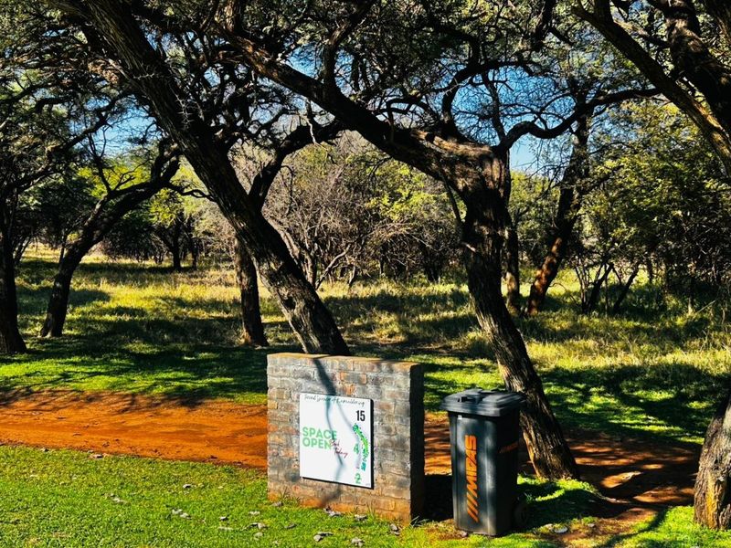 A view of a residential vacant land plot featuring a brick signage pillar and mature trees on a grassed verge.