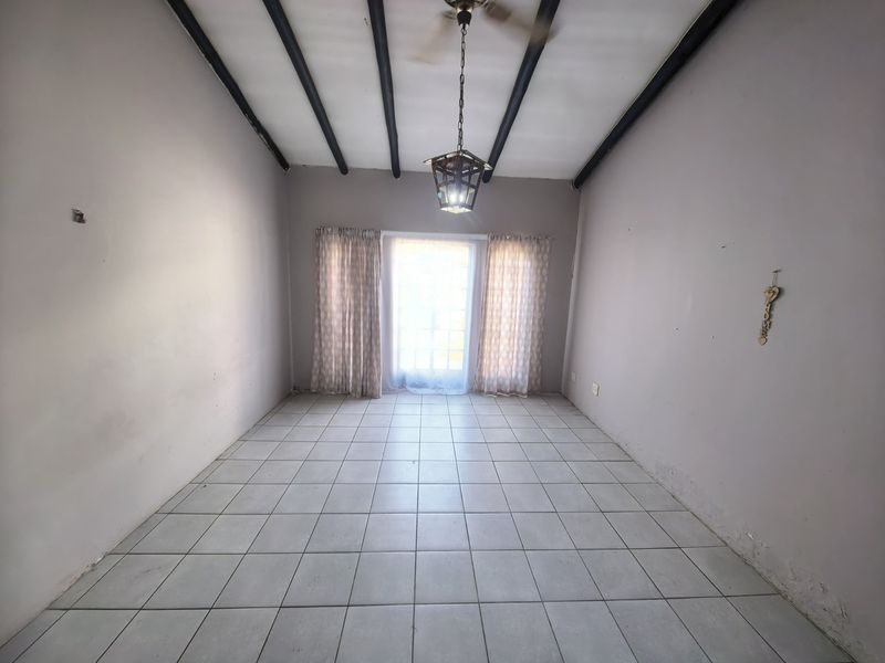 An empty living room featuring tiled flooring, exposed ceiling beams, and a central pendant light, leading to a glass-paned exterior door.
