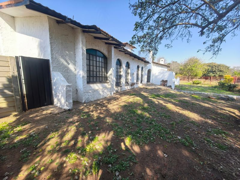 Rear view of a house featuring a textured white facade, arched steel-framed windows, and a tiled roof, overlooking an unlandscaped garden area.
