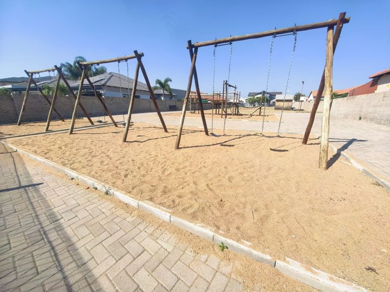 A communal outdoor play area featuring a sand pit, swing sets, and paved pathways, enclosed by a perimeter wall.