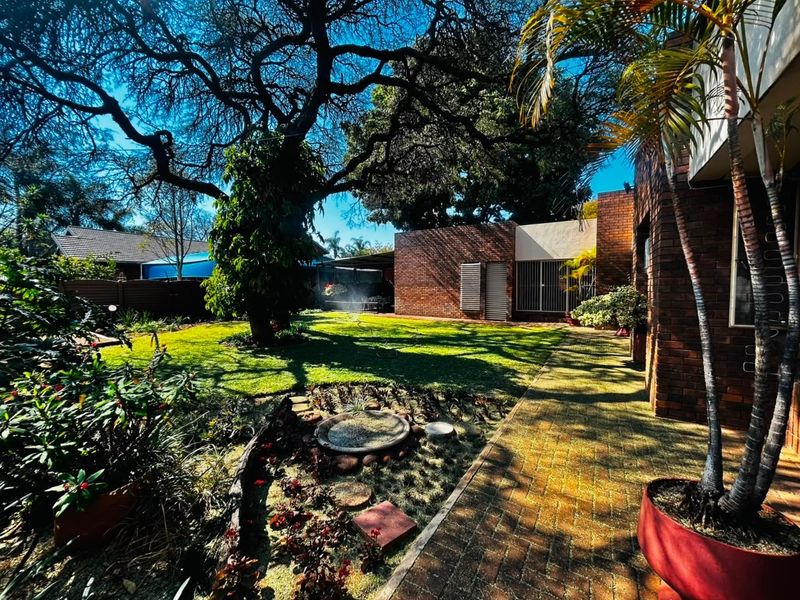 A well-maintained rear garden featuring a brick facade, paved walkway, mature trees, and established landscaping, providing a private outdoor space.