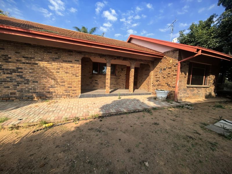 A rear view of a face brick house featuring a covered patio area with brick columns and a paved terrace, set against an unmaintained garden.