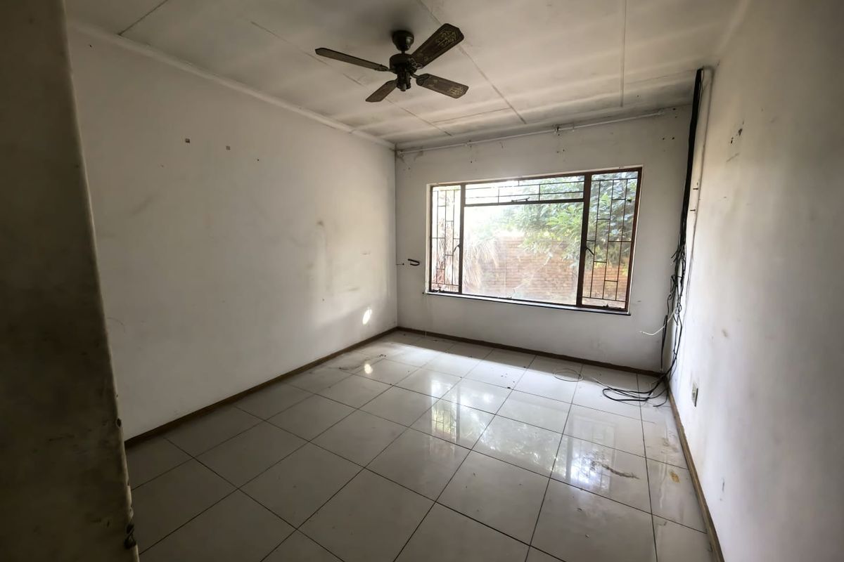 An empty bedroom featuring tiled flooring, a ceiling fan, and a window with security bars, requiring cosmetic updates and repairs to walls and ceiling.