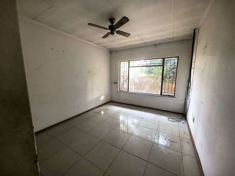 An empty bedroom featuring tiled flooring, a ceiling fan, and a window with security bars, requiring cosmetic updates and repairs to walls and ceiling.