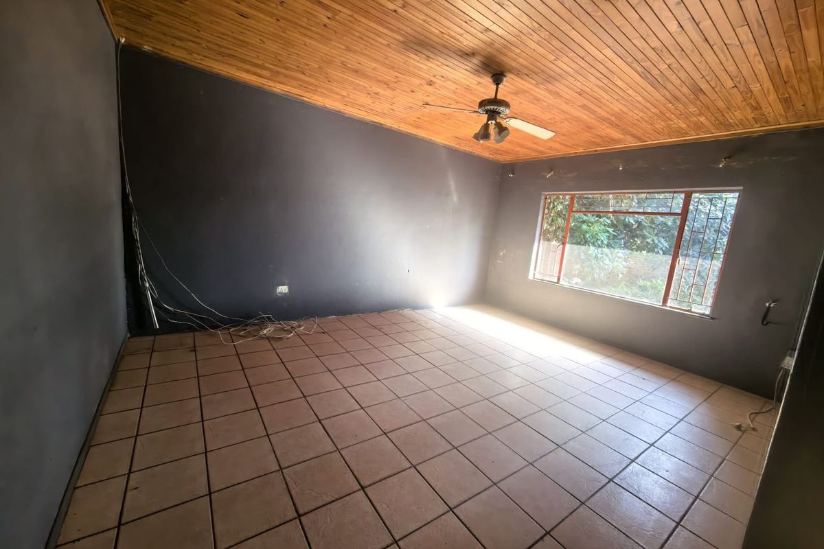 An empty living room featuring a timber-clad ceiling, tiled flooring, and a large window with security bars, requiring minor cosmetic updates.