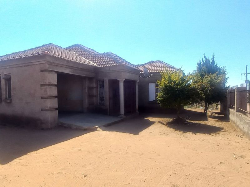 Front view of a single-story house featuring a tiled roof, covered entrance porch, and metal perimeter fencing on a sandy plot.