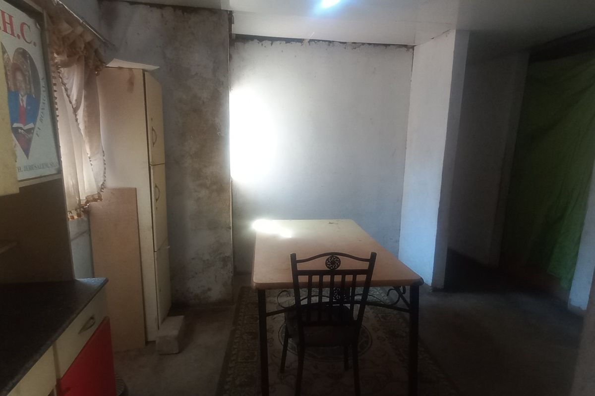 A kitchen area requiring significant renovation, featuring basic cabinetry and showing extensive wall surface deterioration and poor lighting conditions.