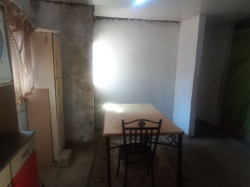 A kitchen area requiring significant renovation, featuring basic cabinetry and showing extensive wall surface deterioration and poor lighting conditions.