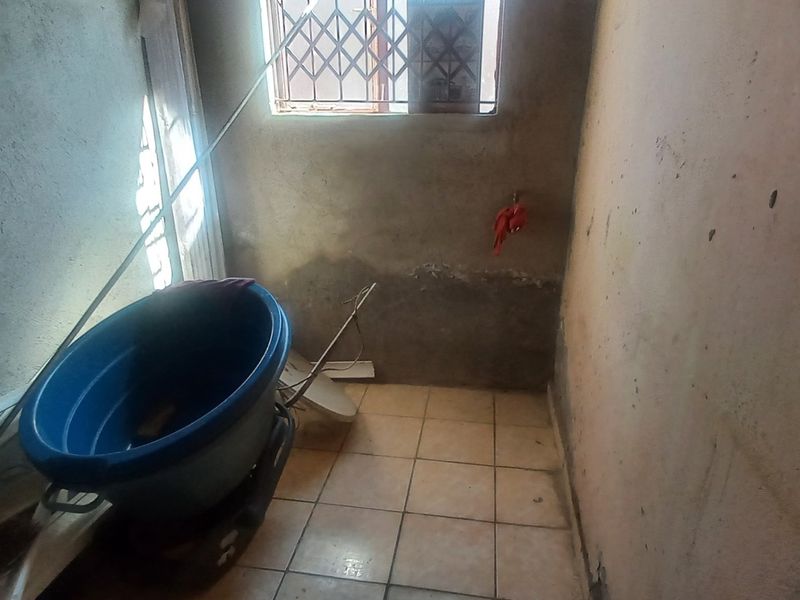 A small, unfinished laundry area featuring tiled flooring and a window with a security gate, currently requiring wall repairs and cosmetic finishing.