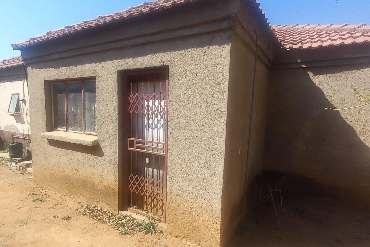 Rear view of a single-story house featuring a tiled roof, security gate, and weathered exterior plaster walls.