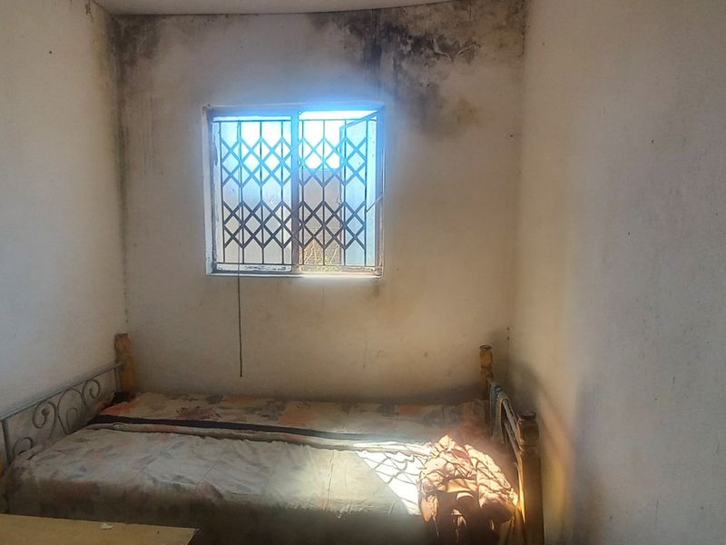 A bedroom featuring a window with a security gate, showing significant signs of wall dampness and deterioration requiring maintenance.