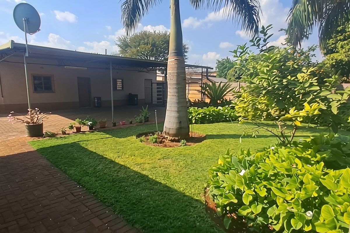 A spacious rear garden featuring a paved patio area, well-maintained lawn, mature palm trees, and a secure boundary wall.