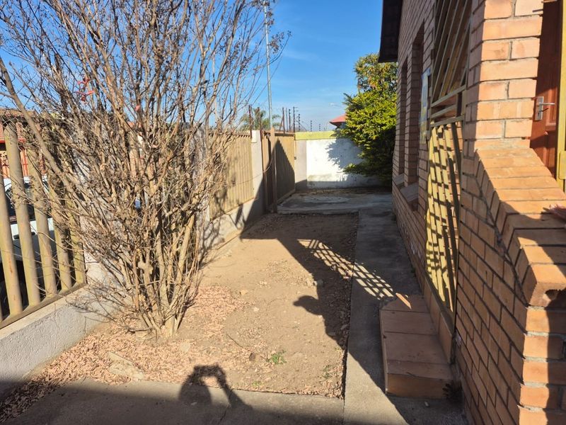 A side exterior view of the property featuring a brick facade, security gate, and a small unlandscaped garden area with concrete paving.
