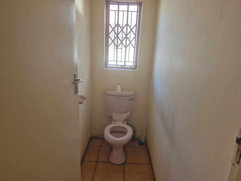 A compact, functional guest toilet featuring tiled flooring and a security-barred window, currently in need of cosmetic updates.