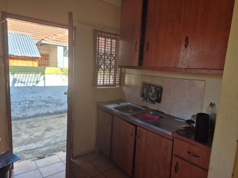 A compact kitchen featuring dated wooden cabinetry, a stainless steel sink, and tiled splashbacks, with direct access to an exterior courtyard area.
