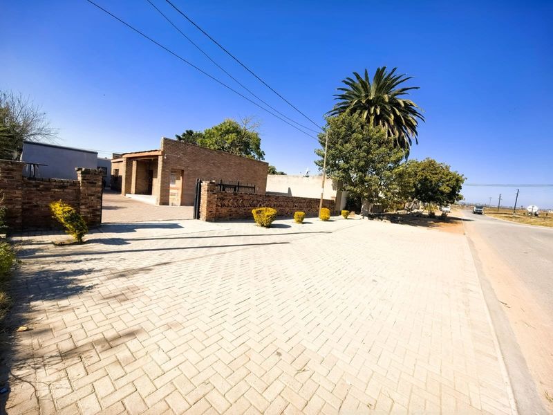 Street view of the property showing the paved driveway, brick boundary wall, and entrance area from the roadside.