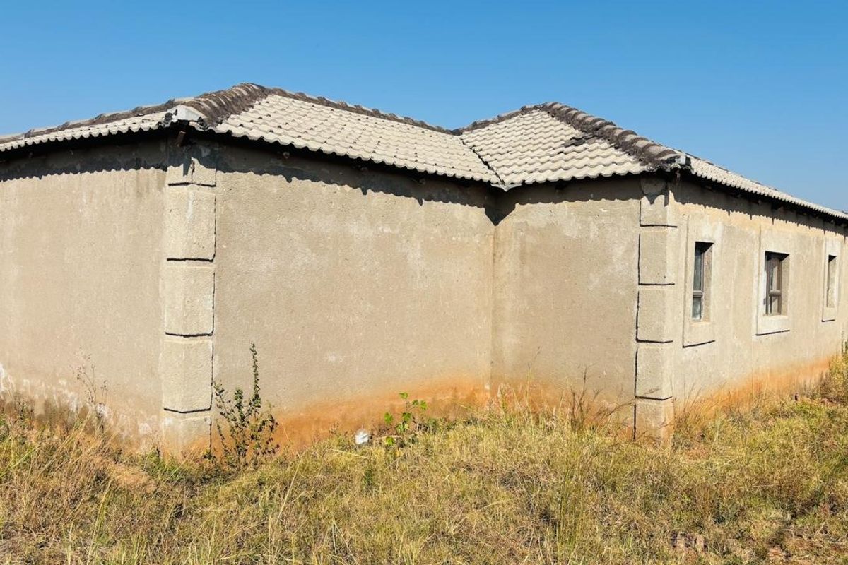 Rear exterior view of a single-story house featuring plastered masonry walls, corner quoins, and a tiled roof, set on an unmaintained plot.