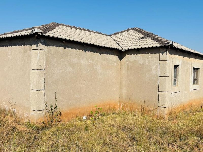 Rear exterior view of a single-story house featuring plastered masonry walls, corner quoins, and a tiled roof, set on an unmaintained plot.