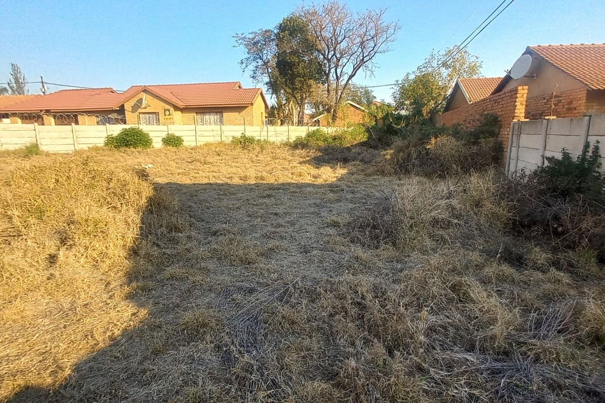 A vacant residential plot enclosed by precast concrete boundary walls with razor wire, featuring overgrown grass and vegetation.