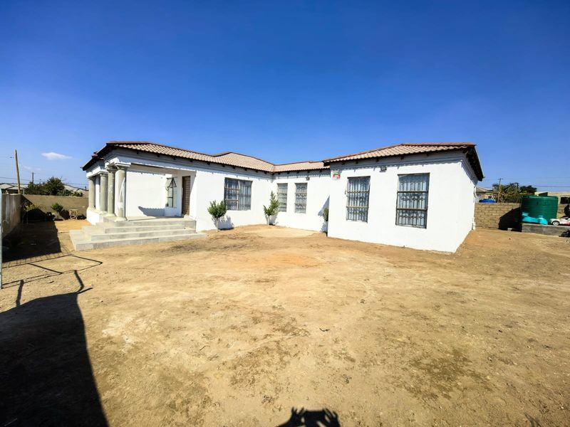 Rear view of a single-story house featuring a white facade, tiled roof, concrete entrance columns, and security bars on windows, set on an unlandscaped plot.