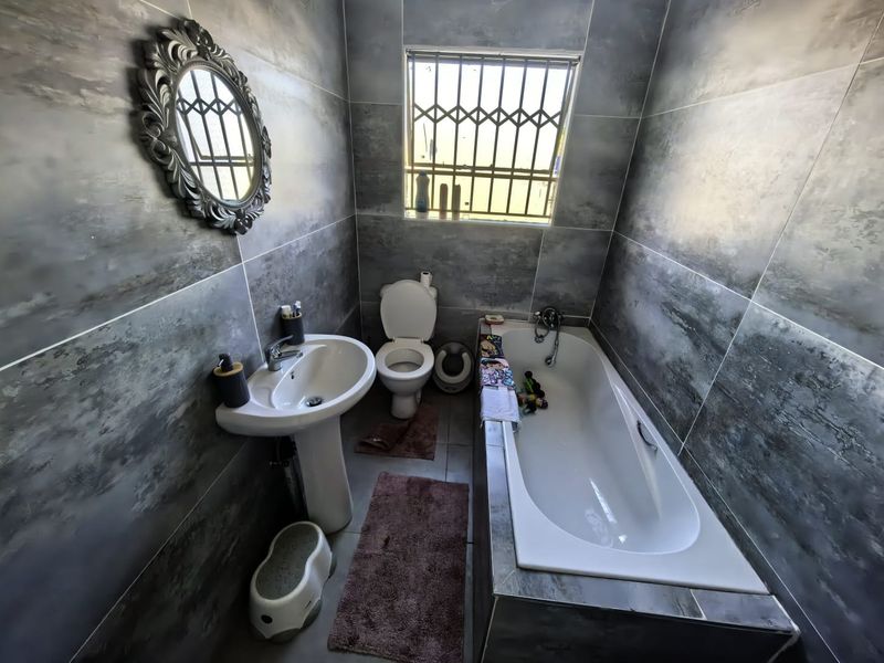 A modern bathroom featuring grey wall tiles, a pedestal basin, a standard toilet, and a built-in bathtub with security bars on the window.