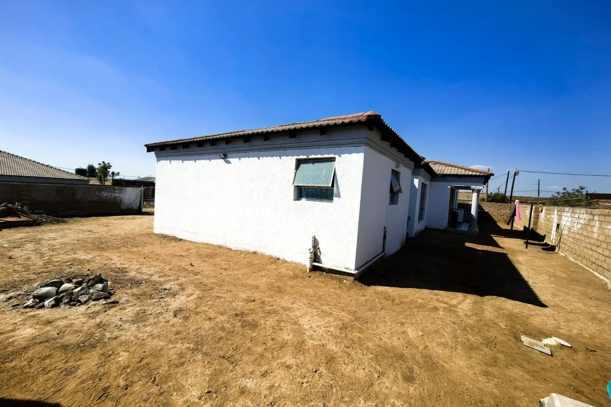 Rear view of a single-story house featuring plastered exterior walls, a tiled roof, and an expansive, unlandscaped yard enclosed by a perimeter boundary wall.