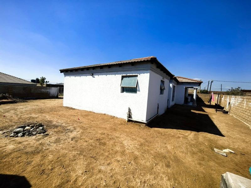 Rear view of a single-story house featuring plastered exterior walls, a tiled roof, and an expansive, unlandscaped yard enclosed by a perimeter boundary wall.
