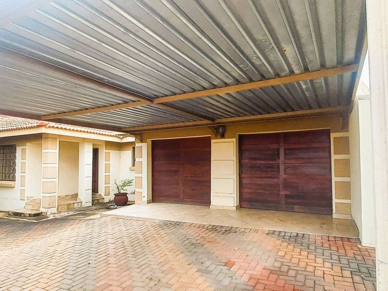 A double garage with wooden doors and a large corrugated metal carport, featuring a paved driveway and established exterior brickwork.
