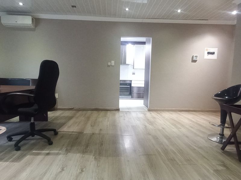 A spacious office area featuring laminate flooring, recessed ceiling lighting, and a wall-mounted air conditioning unit, with a view into the adjacent kitchen.