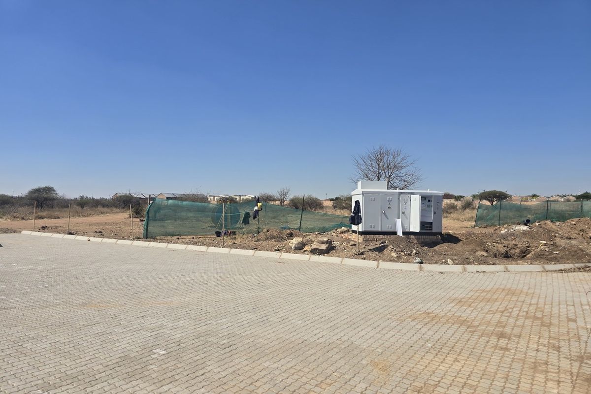 A vacant residential plot featuring a paved driveway, perimeter mesh fencing, and a central electrical substation kiosk on site.