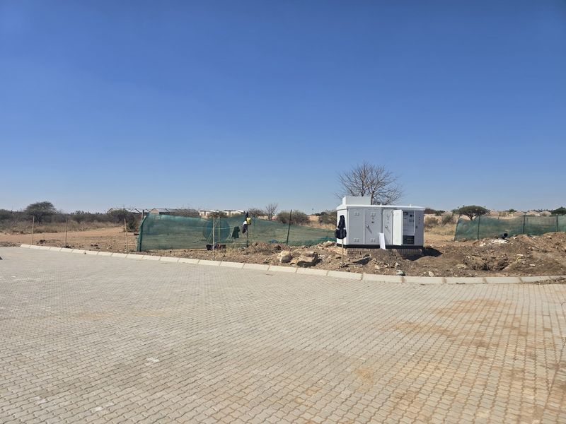A vacant residential plot featuring a paved driveway, perimeter mesh fencing, and a central electrical substation kiosk on site.