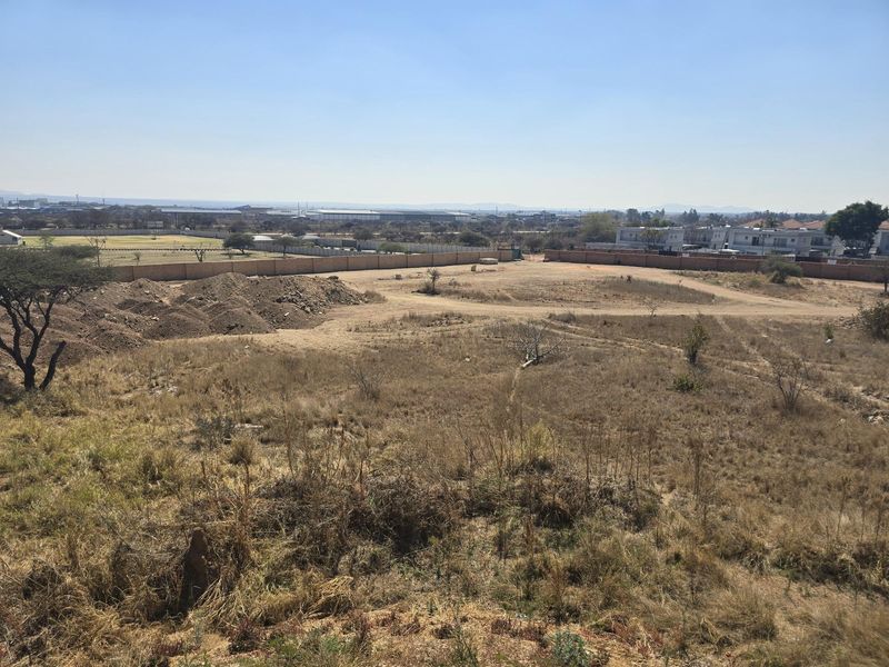 An elevated view of a large, vacant residential plot featuring natural vegetation, perimeter walling, and surrounding neighborhood infrastructure.