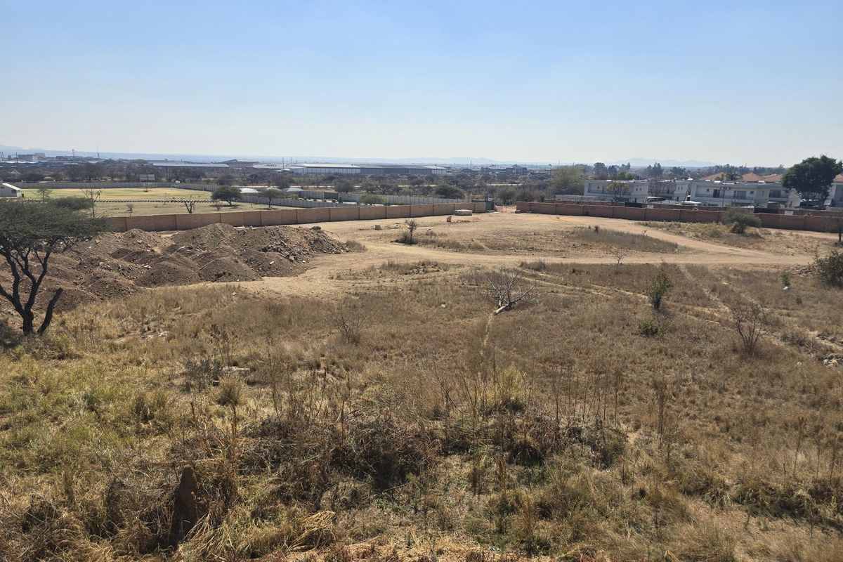 An elevated view of a large, vacant residential plot featuring natural vegetation, perimeter walling, and surrounding neighborhood infrastructure.