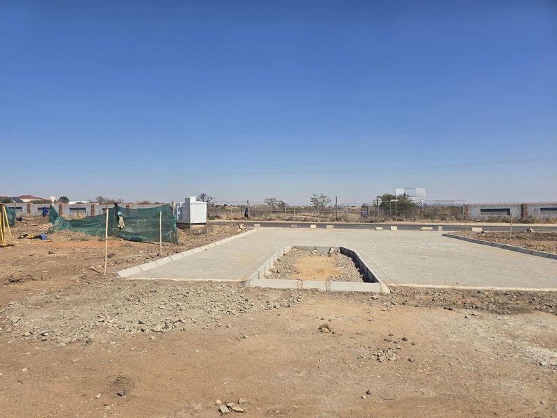 A vacant residential plot featuring a newly installed paved driveway entrance, concrete curbing, and perimeter fencing, ready for future development.