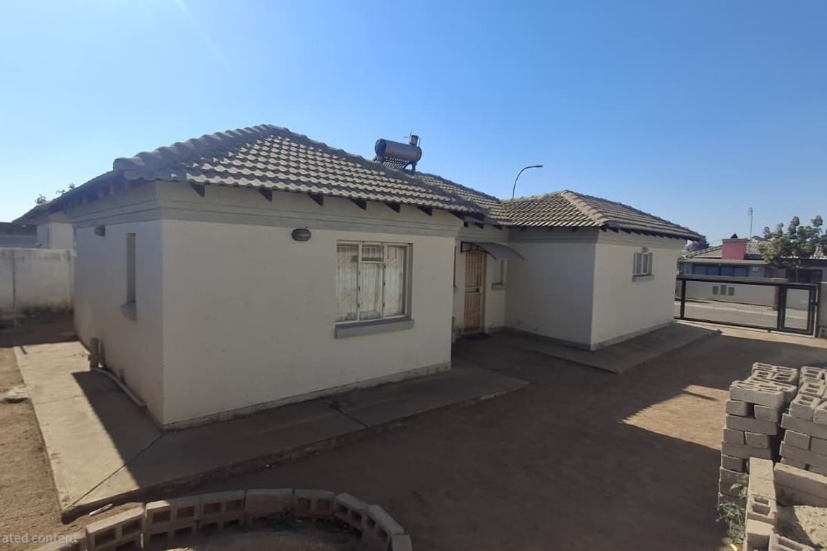 Rear view of a single-story house featuring a tiled roof, solar water heater, and a surrounding concrete perimeter wall with an unpaved yard.