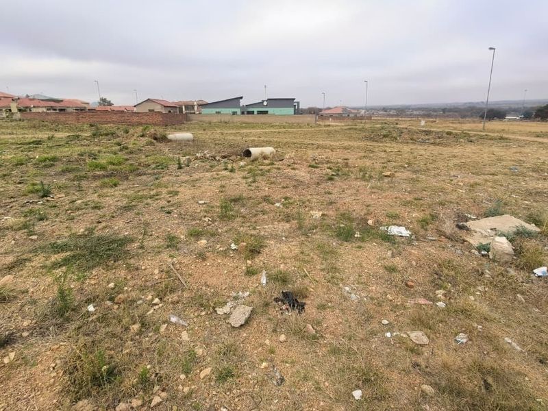 A vacant residential plot of land featuring natural vegetation and scattered concrete drainage pipes, situated in a developing suburban area.